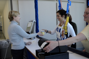 A photo of a woman using the harness system at the Go Baby Go Cafe, handing a female customer a pint of ice cream.
